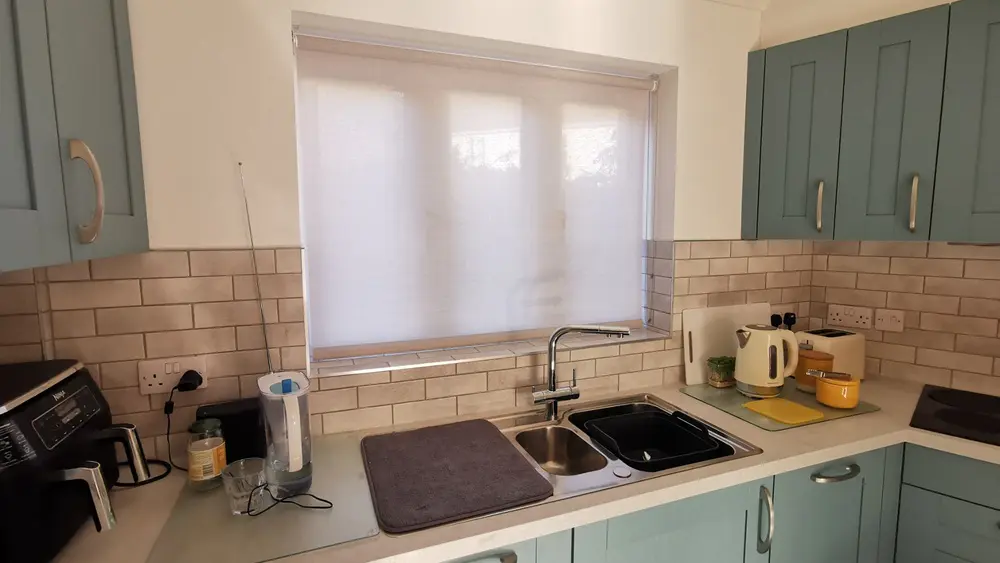 A translucent roller blind fitted above a kitchen sink with teal cabinetry, filtering soft light while cooking